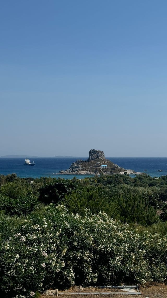 Una vista suggestiva di un isolotto roccioso con un piccolo edificio dal tetto blu in mezzo al mare, incorniciata da cespugli verdi, mentre un traghetto passa sotto un cielo sereno.