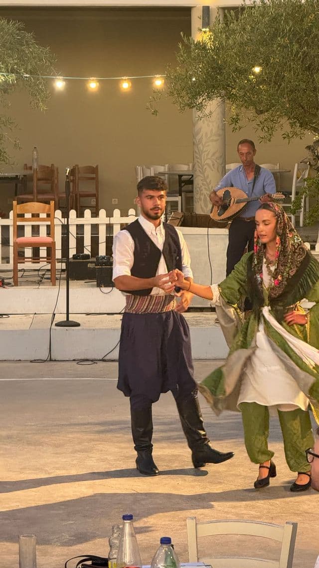 Un uomo e una donna in costumi tradizionali ballano su un palco all'aperto di notte, mentre un musicista suona uno strumento a corde dietro di loro.