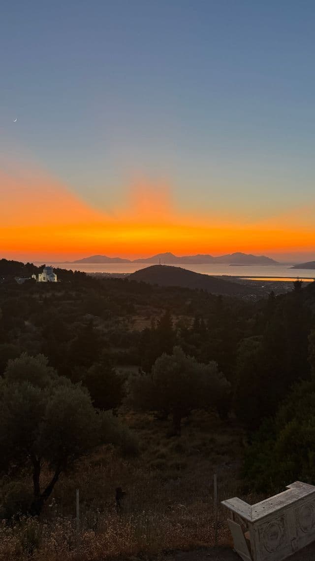 Un vivace tramonto arancione su colline in silhouette e uno specchio d'acqua, con montagne lontane all'orizzonte e una luna crescente nel cielo blu.