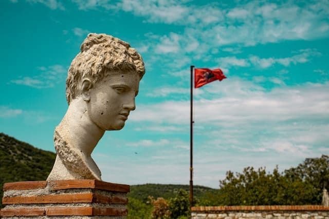 Un busto clásico de piedra de una cabeza descansa sobre un pedestal de ladrillo al aire libre, con la bandera albanesa ondeando al fondo.