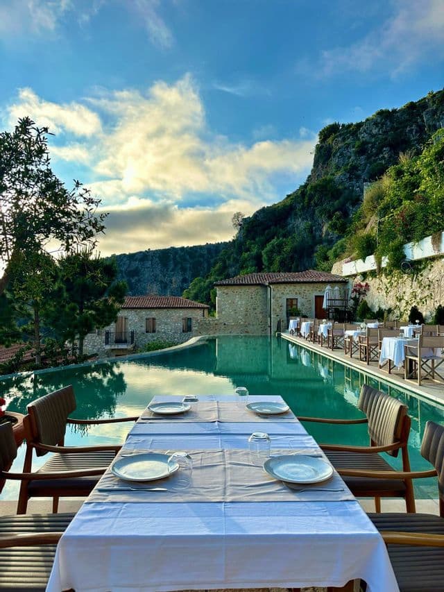 Un restaurante al aire libre con mesas dispuestas a lo largo de una piscina, enclavado en un acantilado rocoso y verde con edificios de piedra.