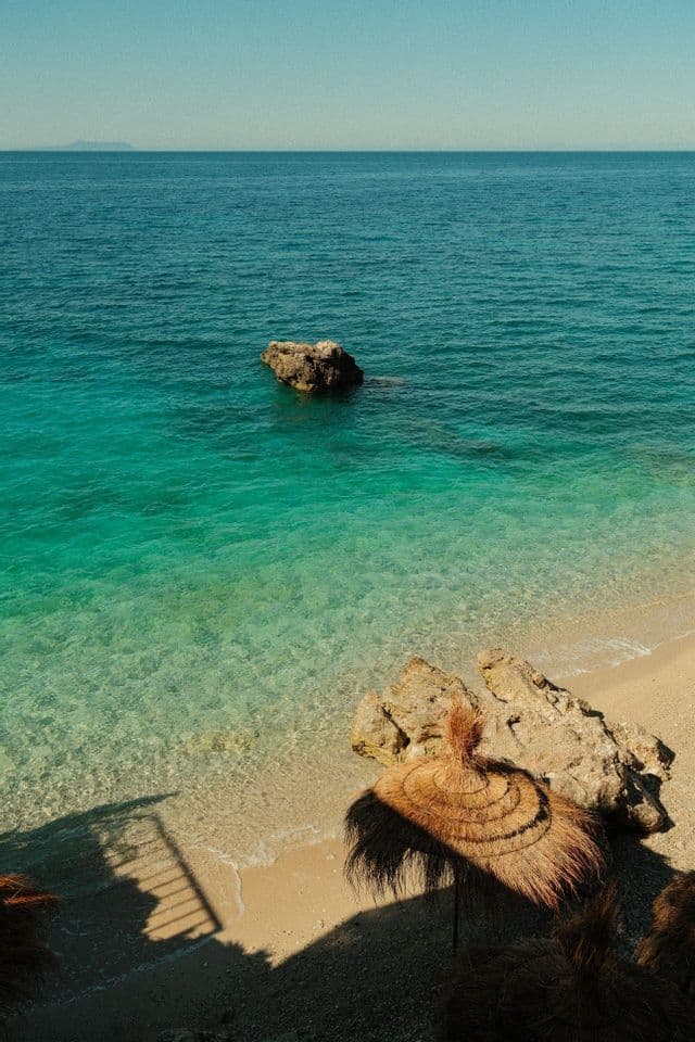 Vista aérea de una sombrilla de playa de paja en una orilla arenosa junto a aguas cristalinas de color turquesa bajo un cielo soleado.
