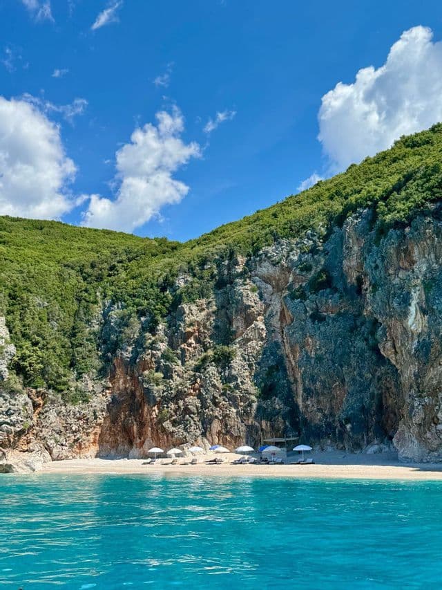 Vista desde el mar turquesa de una playa de arena aislada con sombrillas, al pie de un acantilado rocoso y verde bajo un cielo azul.