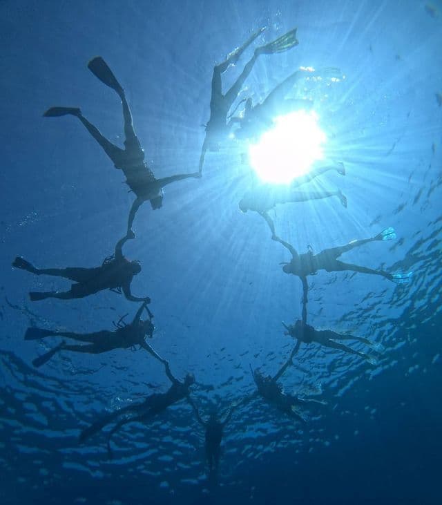 Sott'acqua, un gruppo WeRoad si tiene per mano in cerchio durante lo snorkeling, con le sagome stagliate contro i raggi del sole nel mare blu.