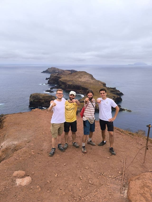 Quattro uomini in viaggio di gruppo WeRoad su una scogliera a picco su una penisola rocciosa e il mare, mentre indicano la fotocamera.