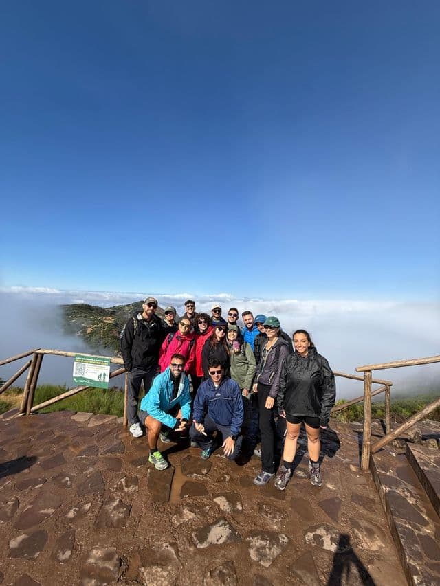 Un gruppo WeRoad in posa per una foto su un punto panoramico in pietra, in alto sopra uno strato di nuvole bianche sotto un cielo blu.