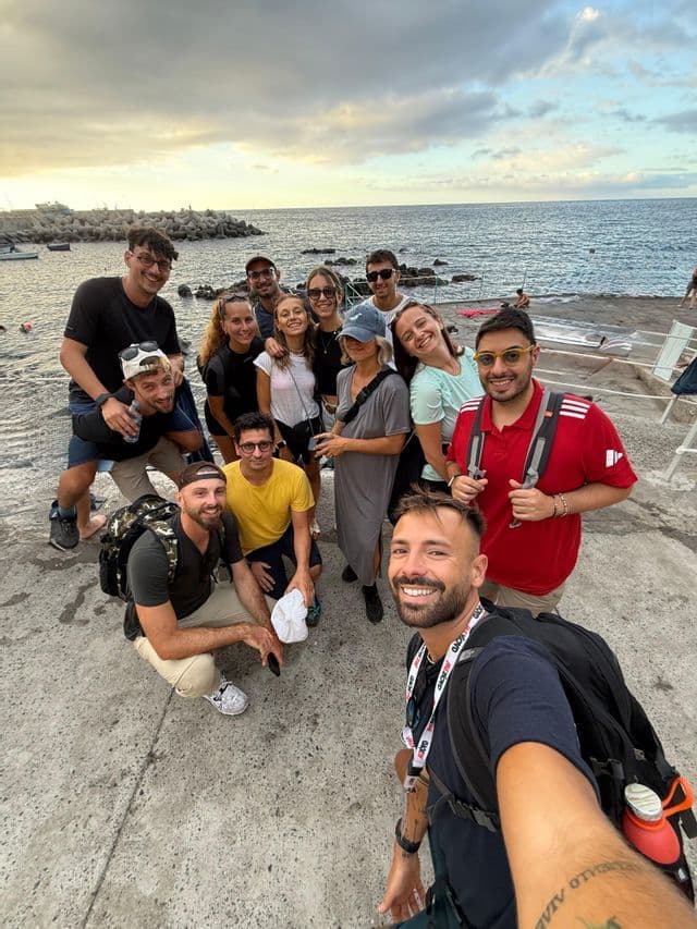 Un gruppo WeRoad si scatta un selfie su un molo di cemento in riva al mare sotto un cielo nuvoloso al tramonto.