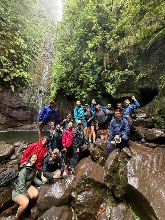 Un gruppo WeRoad in viaggio con impermeabili sorride per una foto su rocce bagnate alla base di una cascata in una foresta rigogliosa.
