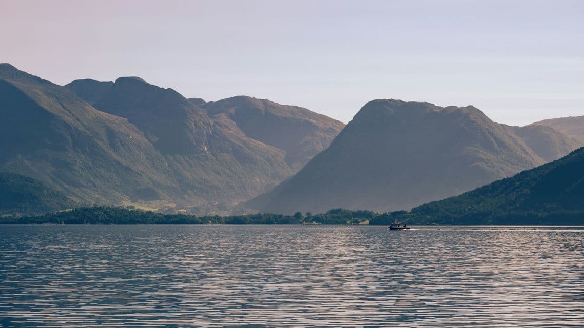 A small boat sails across a calm body of water in front of large, green mountains under a hazy sky.