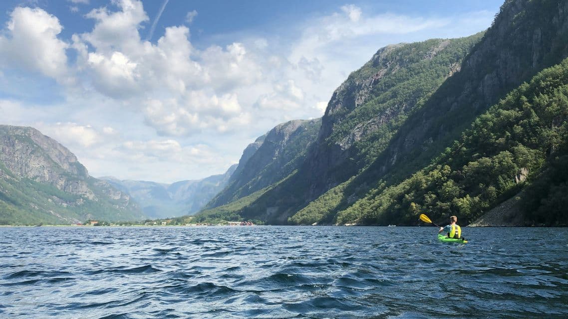 A person in a green kayak paddles on a fjord surrounded by steep, green mountains under a partly cloudy sky.