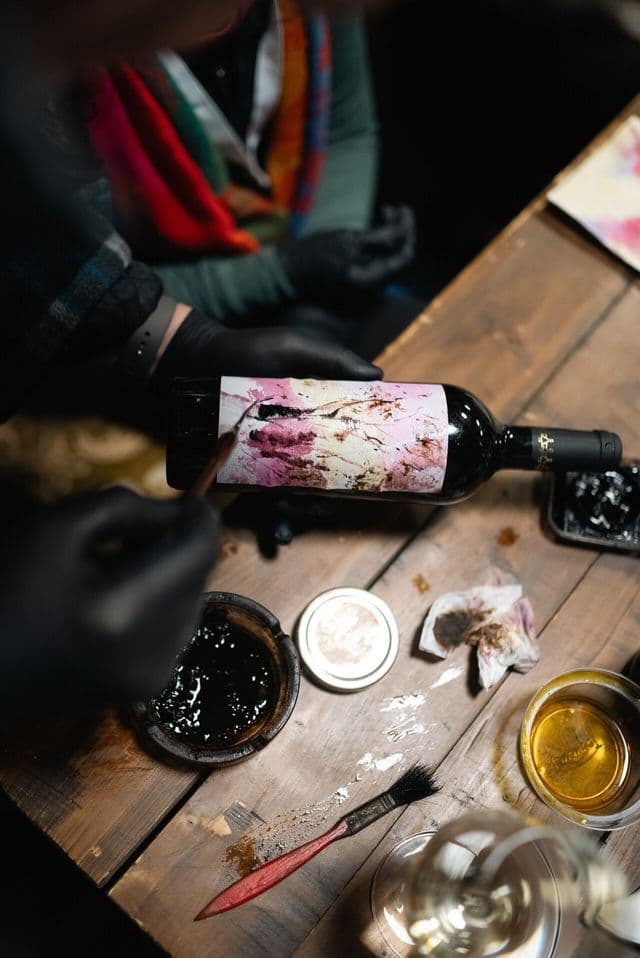 Una persona con guanti neri dipinge un'etichetta personalizzata su una bottiglia di vino rosso, posta su un tavolo di legno con materiali d'arte.