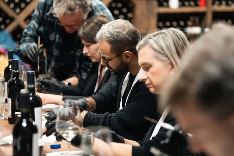 Un gruppo WeRoad con guanti neri partecipa a un workshop in una cantina, a un lungo tavolo con bottiglie e bicchieri di vino.