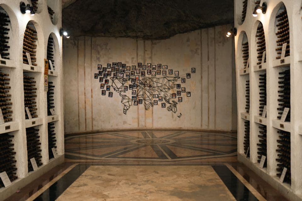 Una cantina con scaffali ad arco pieni di bottiglie e una grande mappa del mondo fatta di foto sulla parete di fondo.