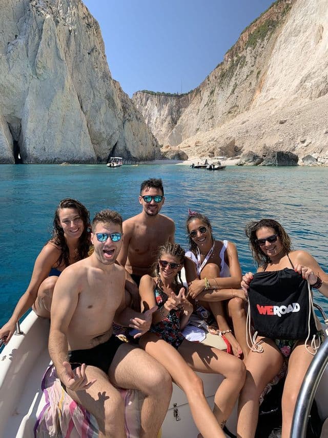 A WeRoad group trip of smiling people posing on a boat in the sea with tall white cliffs in the background.