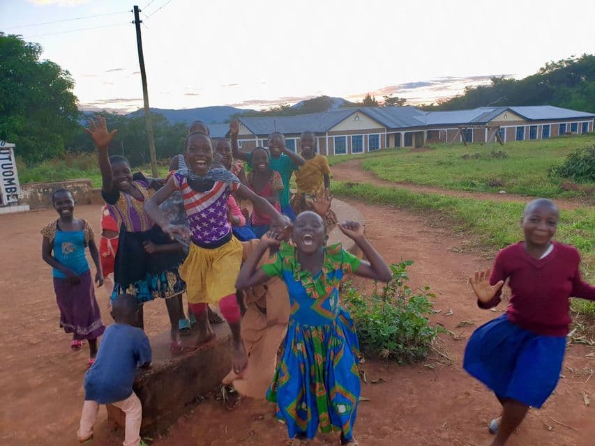 Un gruppo di bambini con vestiti colorati che salutano, ridono e saltano su un sentiero sterrato, con edifici e colline visibili sullo sfondo.