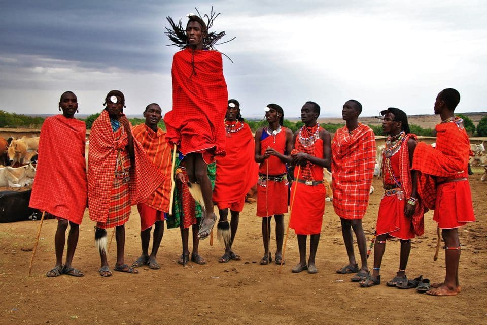Un gruppo di uomini Maasai in tuniche rosse tradizionali sono insieme, mentre uno salta in alto durante una danza tradizionale.