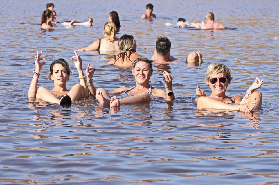 Un viaggio di gruppo WeRoad sorride e galleggia in un lago salato, con tre donne in primo piano che fanno gesti con le mani.