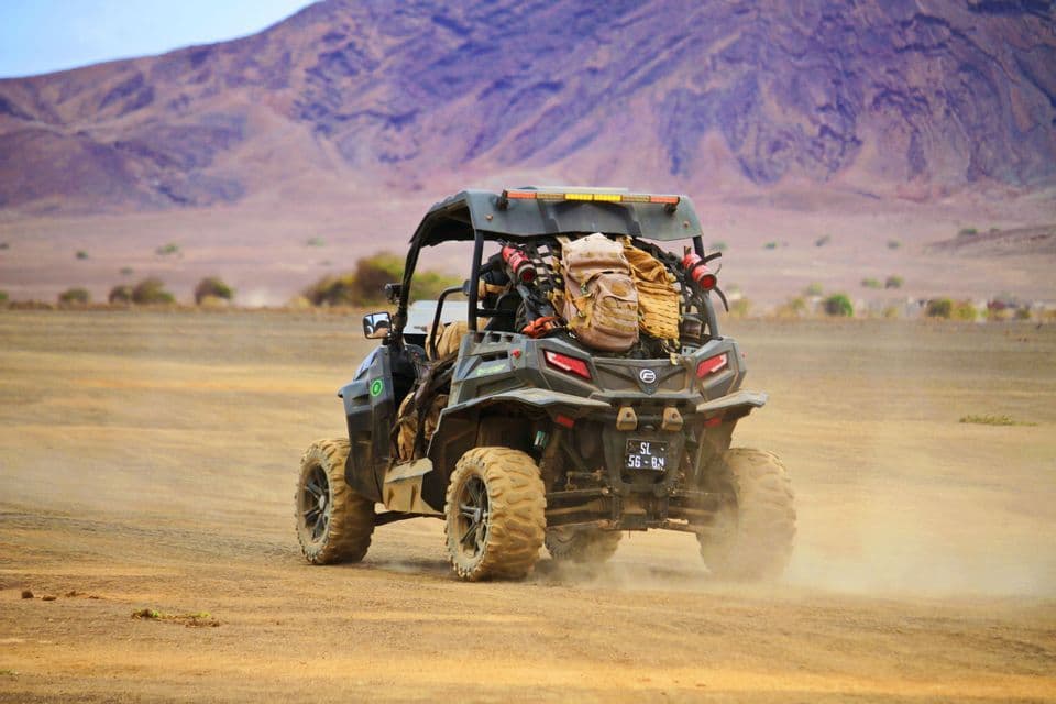 Un viaggio di gruppo WeRoad a bordo di un buggy fuoristrada attraversa un paesaggio desertico polveroso con montagne sullo sfondo.