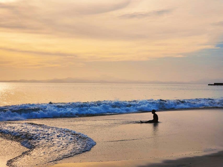 Una persona en silueta sentada en una playa de arena mojada, con una ola acercándose y bajo un cielo de atardecer dorado.