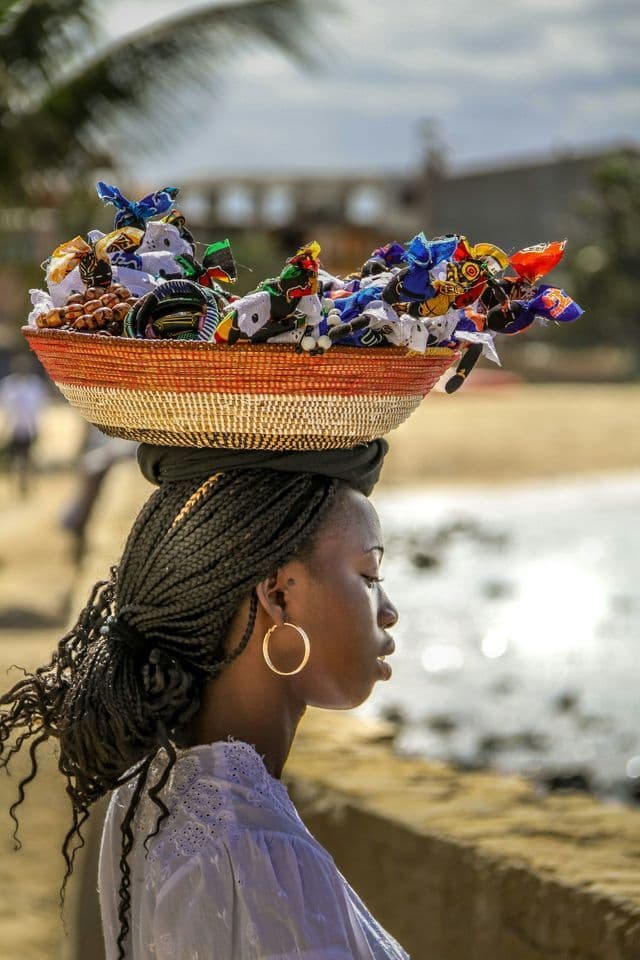 Una mujer con trenzas equilibra una canasta tejida llena de artesanías coloridas sobre su cabeza, cerca de una playa.