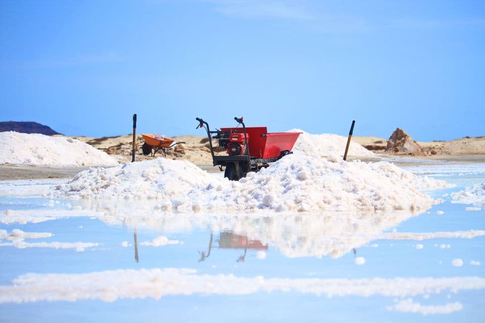 Una carretilla motorizada roja se encuentra entre montones de sal blanca en un salar, con su reflejo visible en el agua en primer plano.