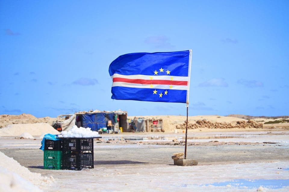 La bandera de Cabo Verde ondea al viento sobre un vasto salar con pequeños edificios en la distancia bajo un cielo azul.