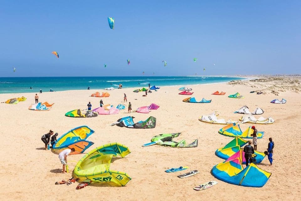 Una spiaggia soleggiata piena di gente che prepara colorate attrezzature da kitesurf, con molti aquiloni già in volo sull'oceano.