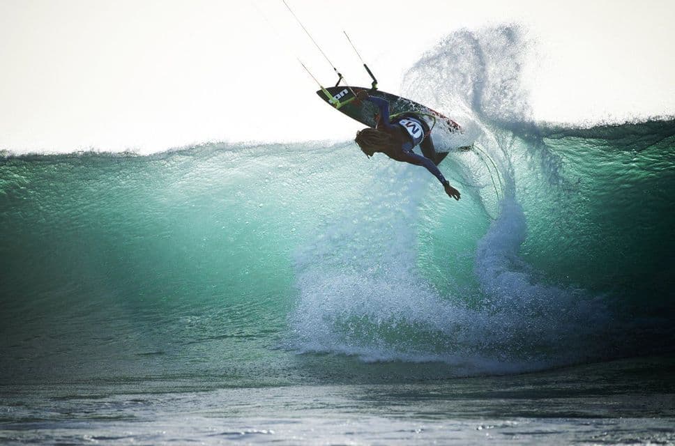 Un kitesurfer con i capelli lunghi cavalca la cresta di una grande onda turchese, sollevando uno spruzzo d'acqua.