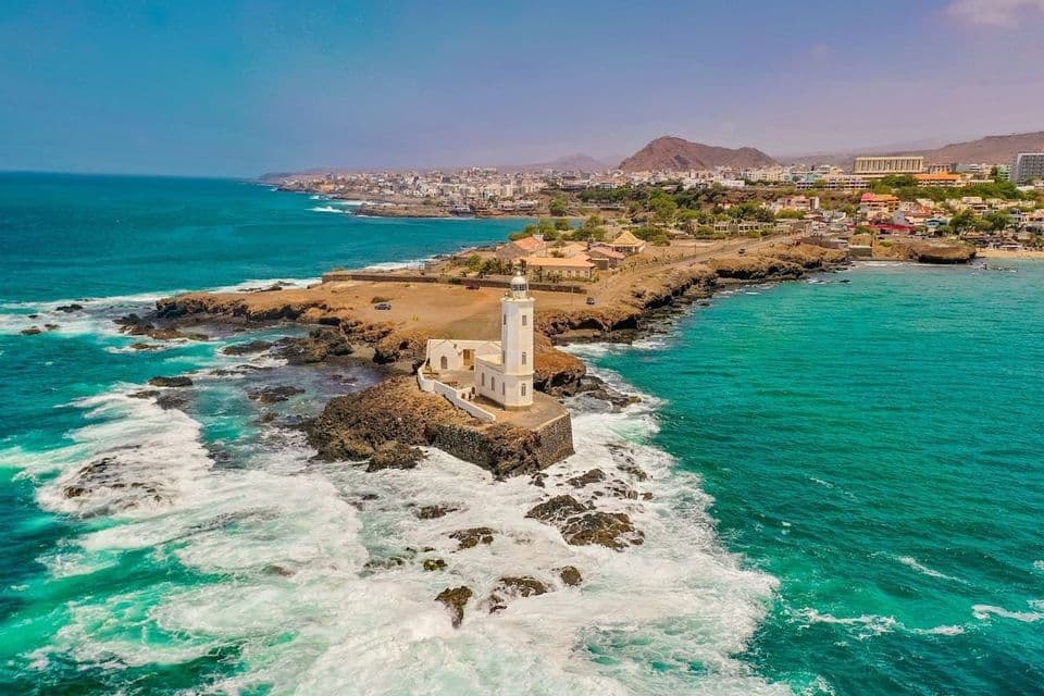 Una vista aerea di un faro bianco su un isolotto roccioso, con onde turchesi che si infrangono sulla riva e una città sullo sfondo.