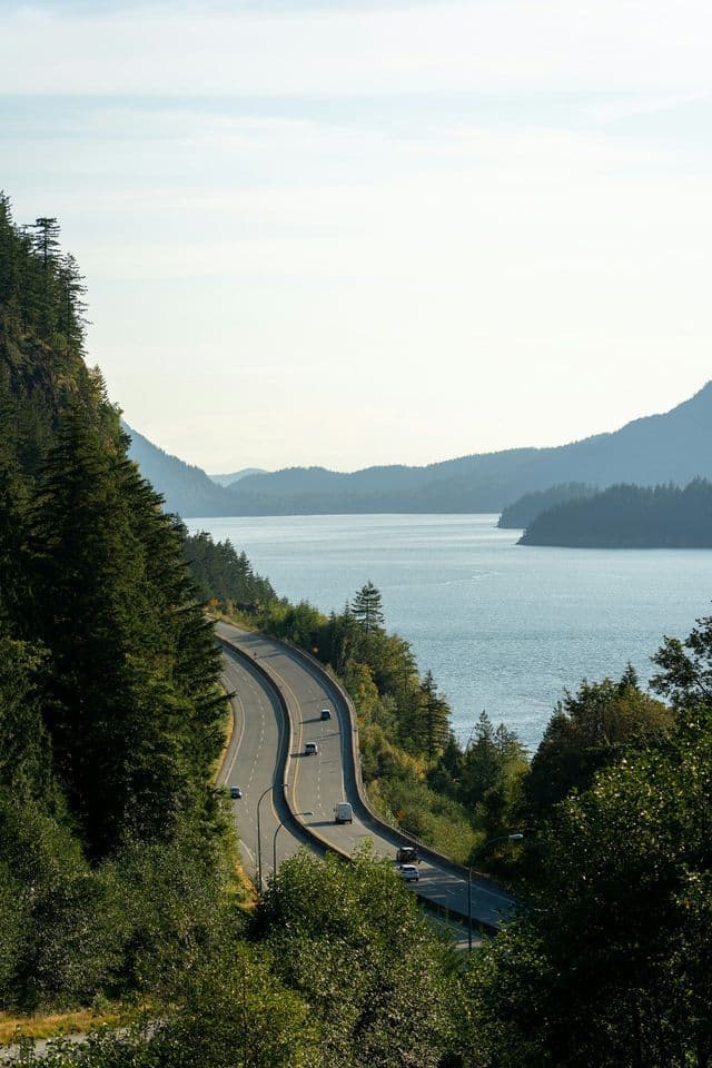 Una vista aerea di un'autostrada tortuosa con auto che guidano lungo una scogliera boscosa accanto a un grande specchio d'acqua e montagne.