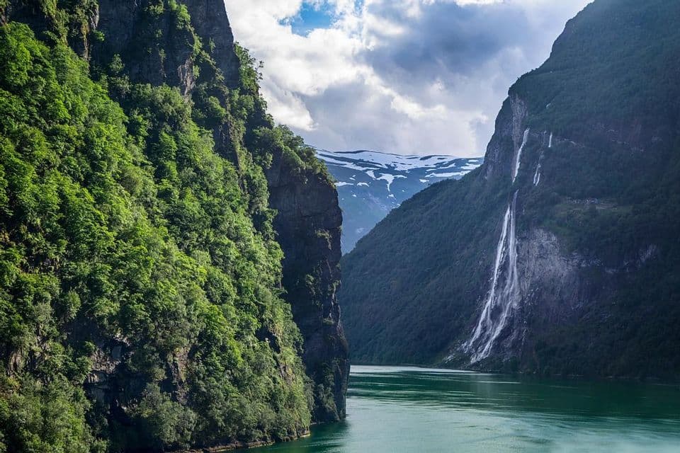 A waterfall cascades down a steep, green mountainside into a fjord, with a snow-capped mountain peak visible in the distance.