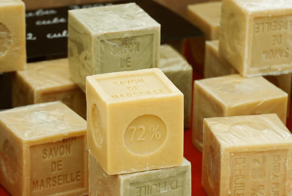 A close-up of stacked, cube-shaped bars of Savon de Marseille soap. One unwrapped bar in the foreground is stamped with '72%'.