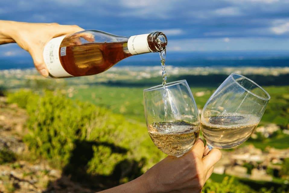 A person pours rosé wine from a bottle into two glasses overlooking a lush green valley.