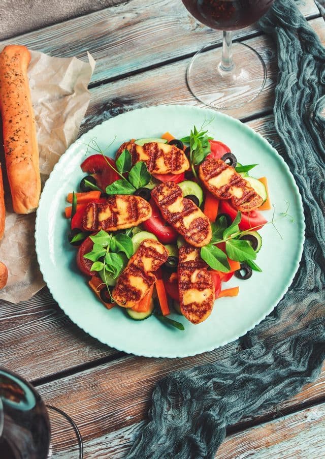 Un piatto di insalata con formaggio halloumi grigliato, accompagnato da grissini e un bicchiere di vino rosso su un tavolo di legno.
