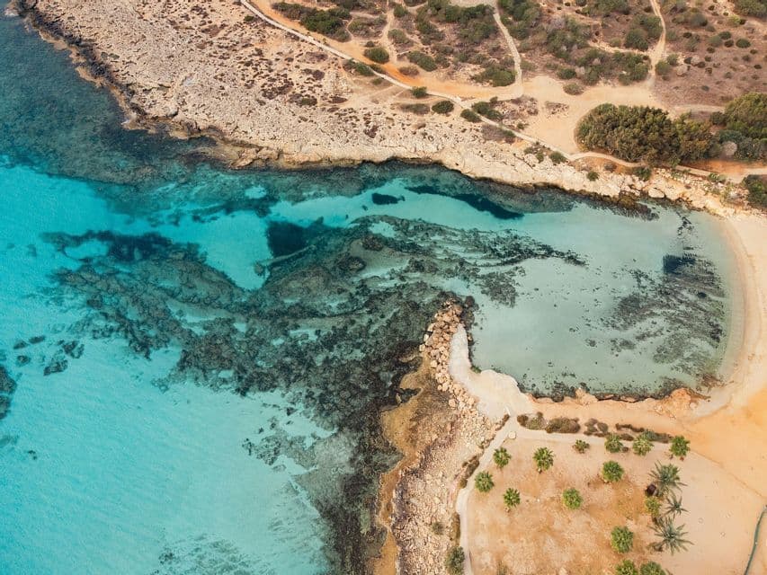 Una veduta aerea di una costa rocciosa accanto ad acque turchesi limpide e una piccola spiaggia sabbiosa con palme.