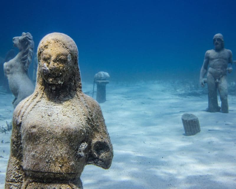 Una scena sottomarina che mostra diverse statue di pietra consumate dal tempo adagiate su un fondale oceanico sabbioso.