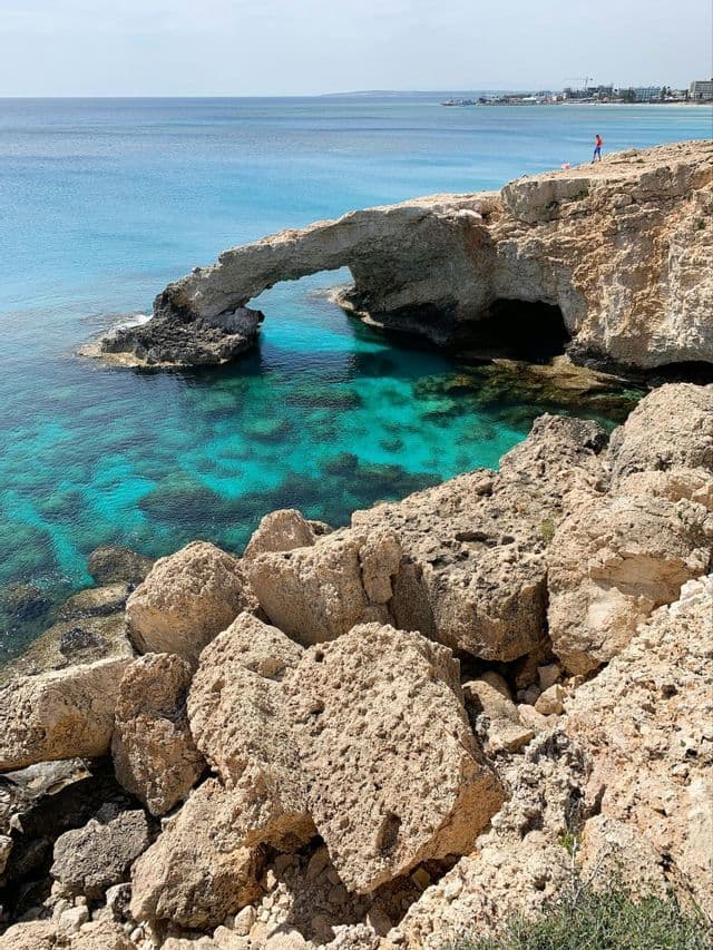 Un arco di roccia naturale su una costa frastagliata si protende su acque limpide e turchesi sotto un cielo soleggiato.