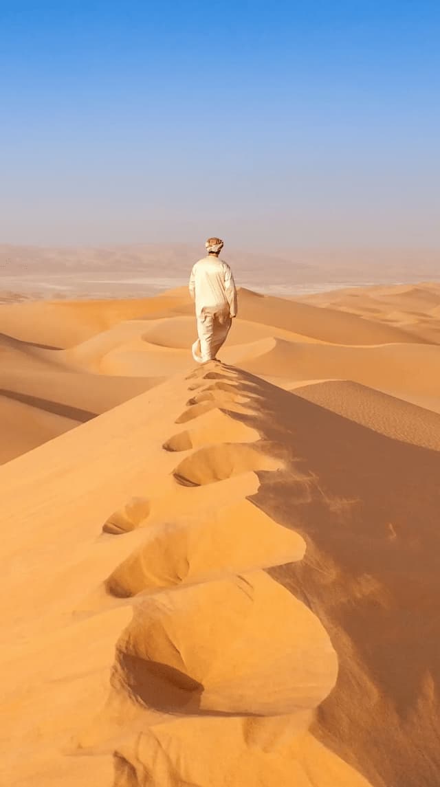 A person in traditional attire walks along the crest of a large sand dune in a vast desert, leaving footprints behind them.