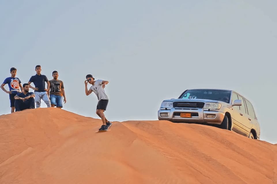 A man from a WeRoad group trip sandboards down a large desert dune as his friends watch from the top, next to a silver SUV.