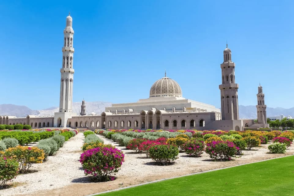 A large, light-colored mosque with a patterned dome and tall minarets, set behind a manicured garden with rows of colorful flowering bushes.
