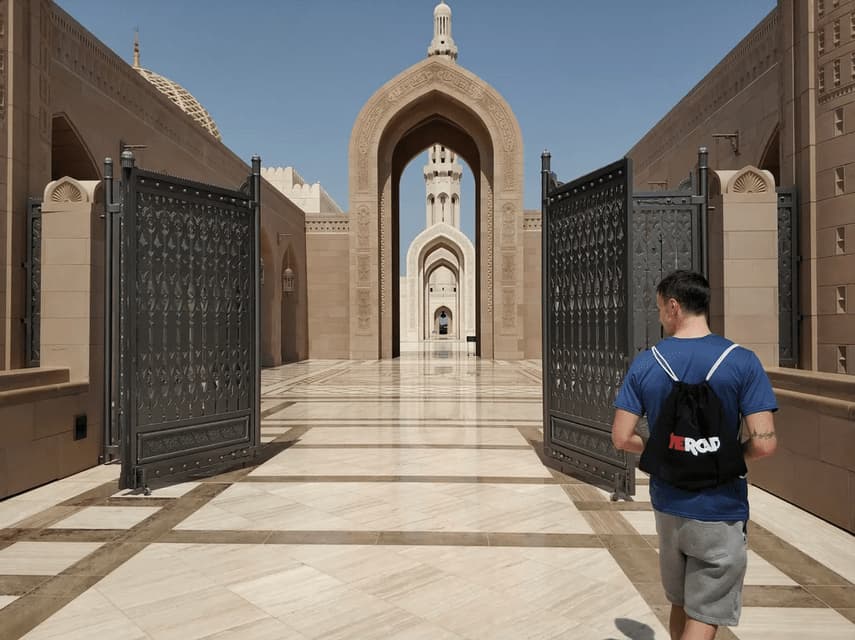 A man wearing a WeRoad backpack walks from behind through an open gate towards a large stone building with arches.