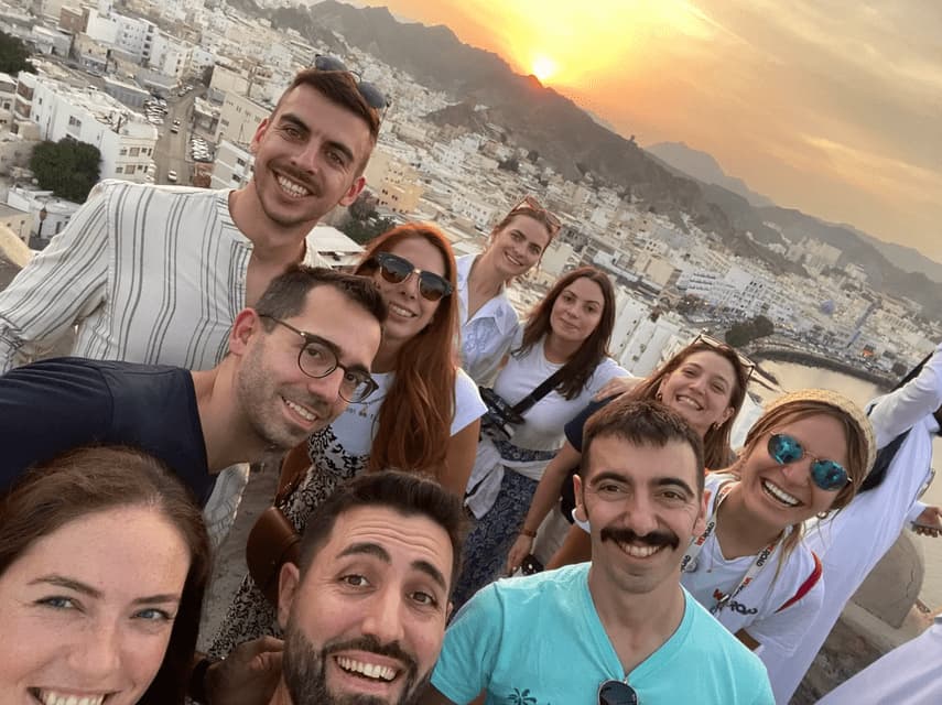 A WeRoad group trip takes a smiling selfie overlooking a city with white buildings as the sun sets behind the mountains.