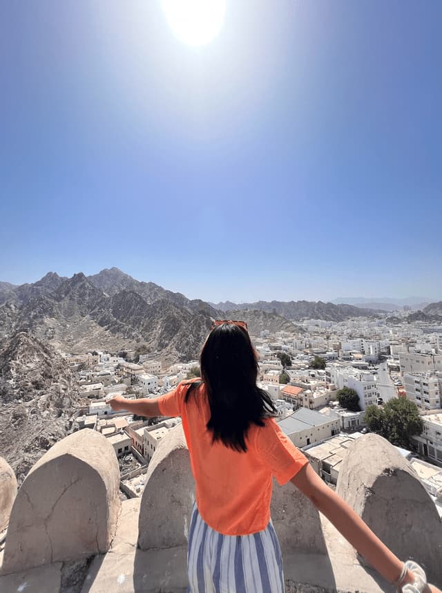 A woman in an orange shirt stands on a stone viewpoint with her arms outstretched, overlooking a city nestled in rocky mountains.