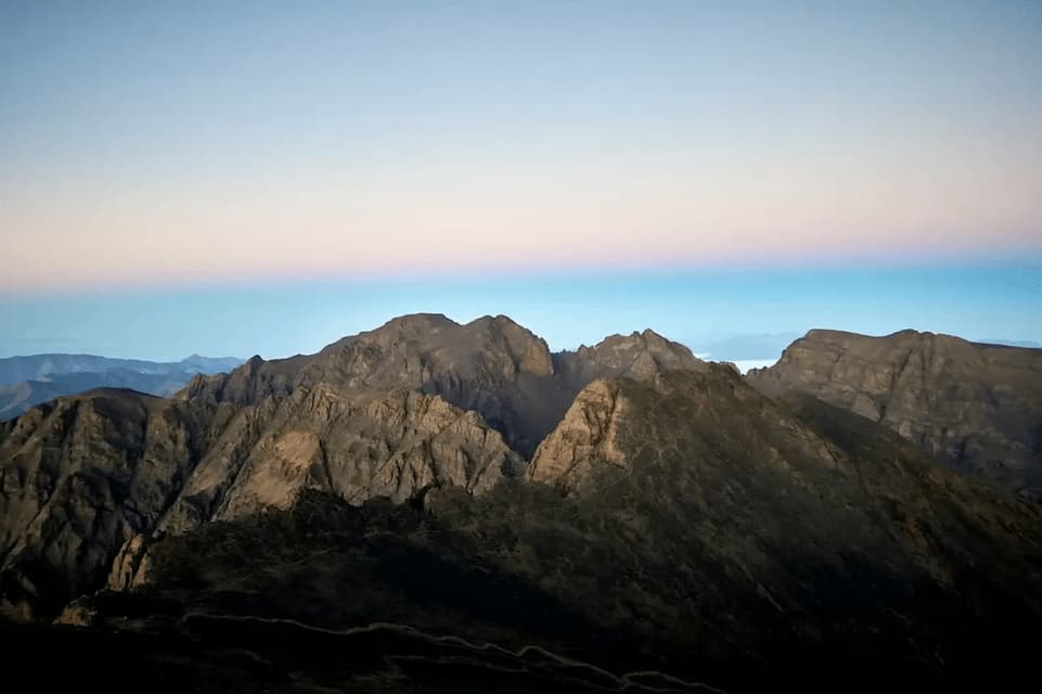 Felsige Berggipfel stehen vor einem klaren Himmel mit blau-rosafarbenen Lichtbändern bei Sonnenaufgang oder Dämmerung.