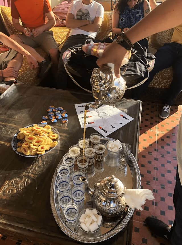 Eine Person gießt traditionellen Tee aus einer silbernen Teekanne in Gläser für eine WeRoad Gruppenreise, die sich um einen Tisch mit Keksen versammelt hat.