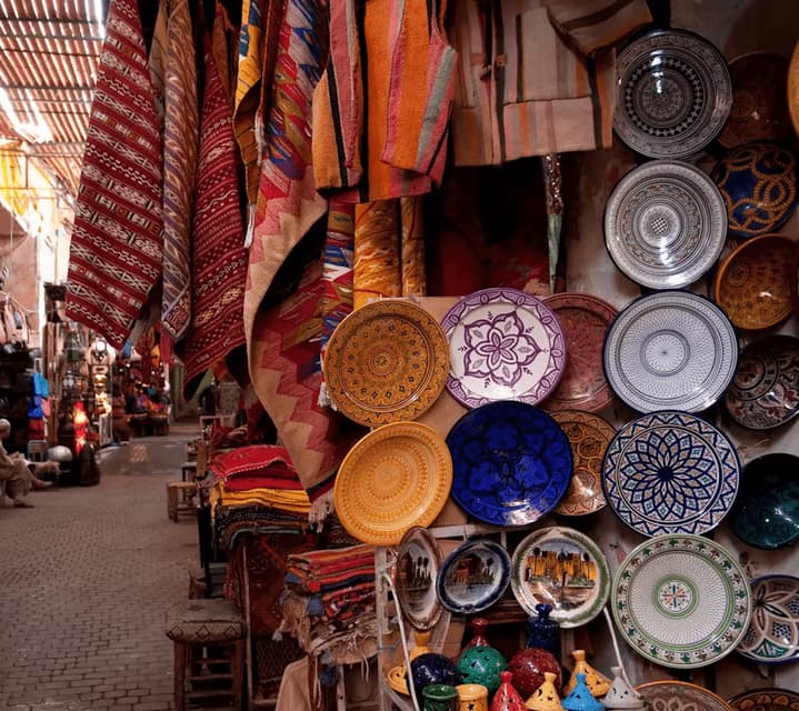 Un étal de marché expose des assiettes en céramique colorées et à motifs et des textiles suspendus le long d'une étroite ruelle pavée.