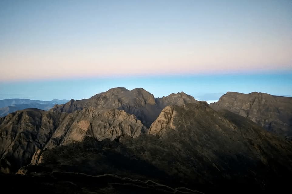 Schroffe, dunkle Berggipfel ragen vor einem klaren Himmel mit einem rosa-blauen Farbverlauf am Horizont im Morgengrauen.