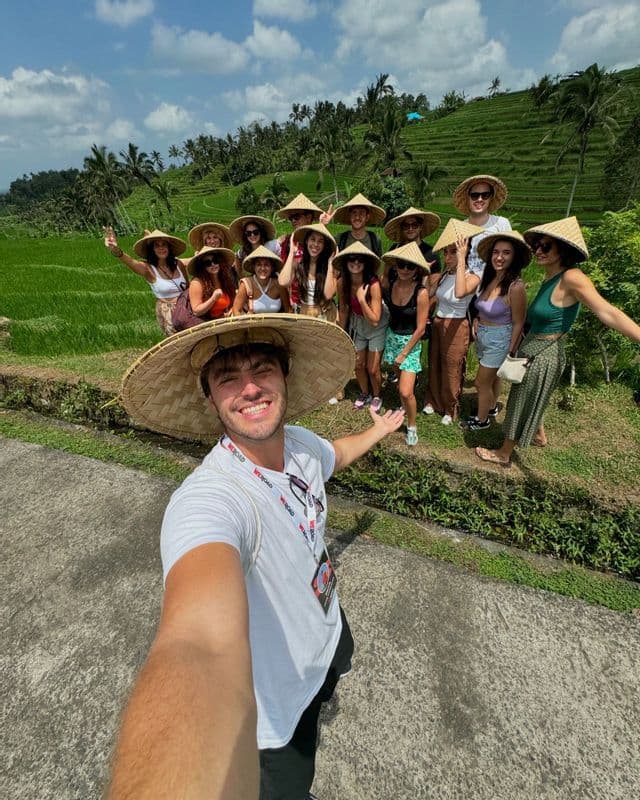 Un uomo si fa un selfie con un gruppo WeRoad, tutti con cappelli di paglia conici, davanti a verdi risaie a terrazze.