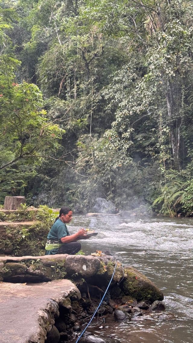 Una donna si inginocchia su una sponda rocciosa accanto a un fiume che scorre, tenendo un vassoio fumante per offerte durante un rituale in una foresta lussureggiante.