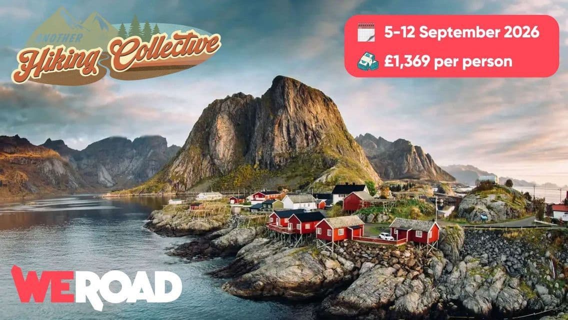A coastal village with red houses on stilts sits on a rocky shore beside calm water at the base of a large mountain.